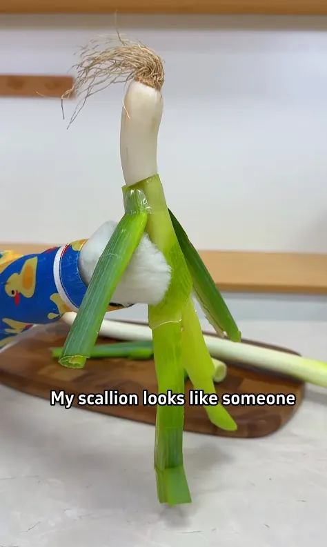 A scallion arranged to look like a tiny standing figure on a wooden board, held up beside Puff's duck-pajama sleeve.