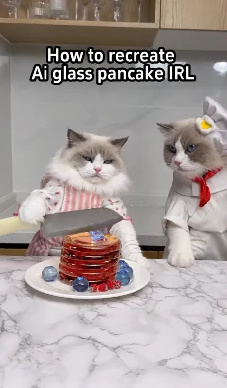 Two cats in aprons at a marble counter slicing a stack of translucent red pancakes topped with glass-like marbles.